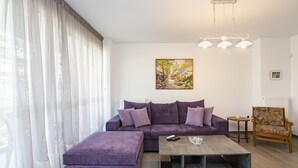 A modern living room with a purple sofa, a matching ottoman, a coffee table, and a patterned armchair. A painting hangs on the wall, and a chandelier is visible. - Beautiful flat in superb location (Glyfada)