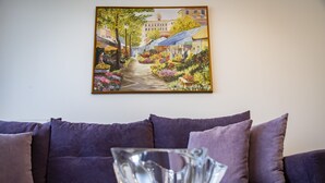 A living room with a purple sofa, a glass bowl on a table, and a painting of a street scene on the wall. - Beautiful flat in superb location (Glyfada)