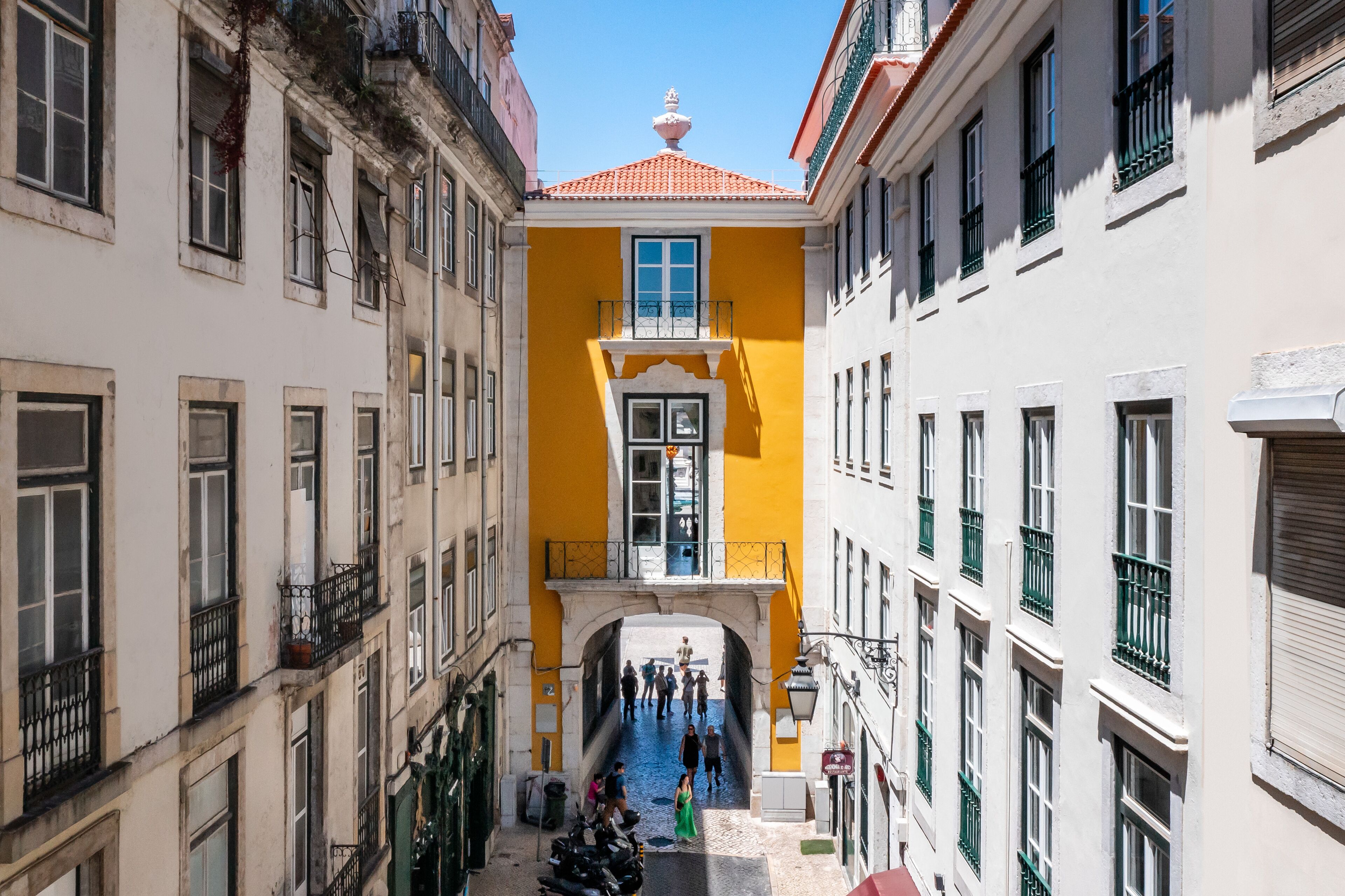 Una calle estrecha flanqueada por edificios, con un edificio central amarillo y un balcón, y un cielo azul despejado.