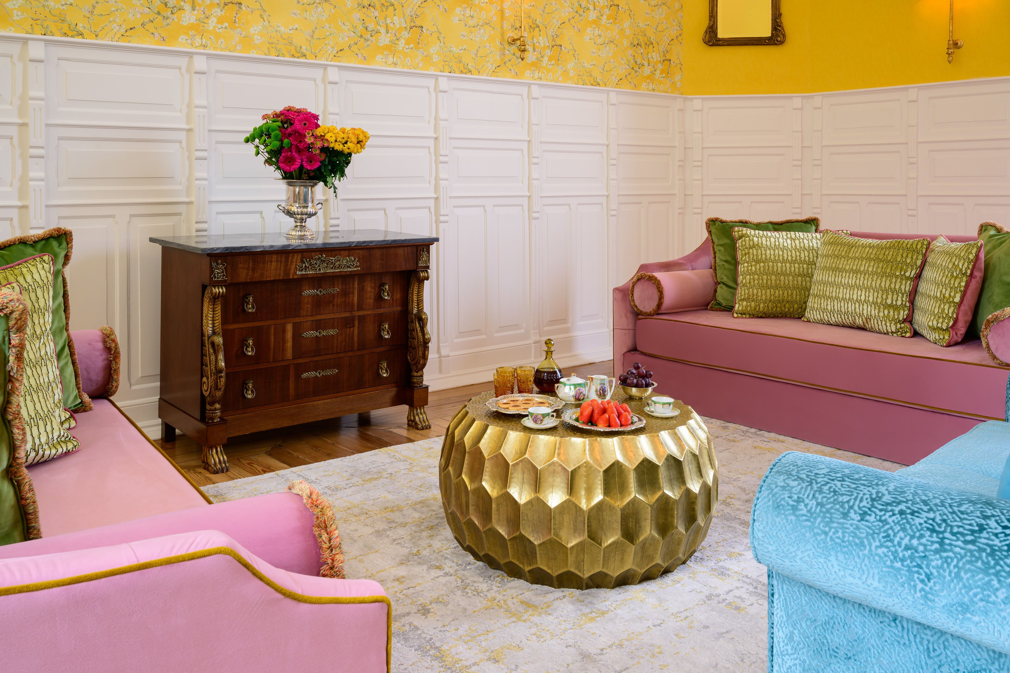 A room with a wooden dresser, a round gold coffee table, a pink sofa, and a blue armchair.