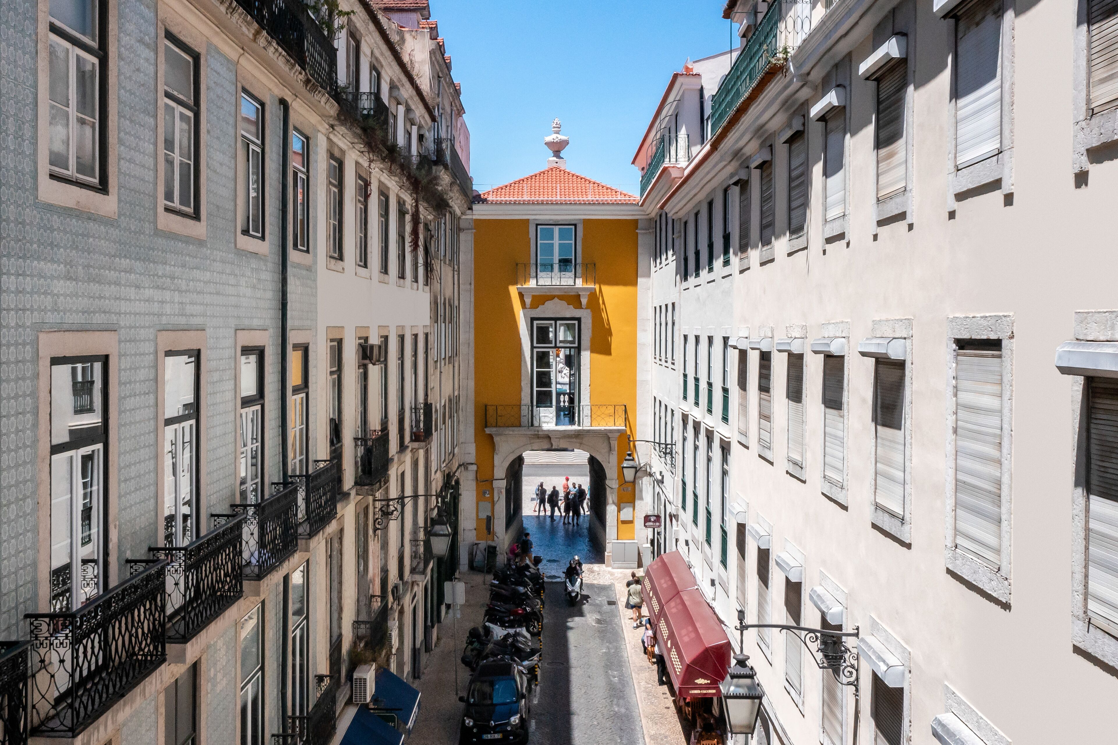 Eine schmale Straße mit Gebäuden an beiden Seiten, ein gelbes Gebäude mit rotem Dach und ein weißes Gebäude mit Balkon.
