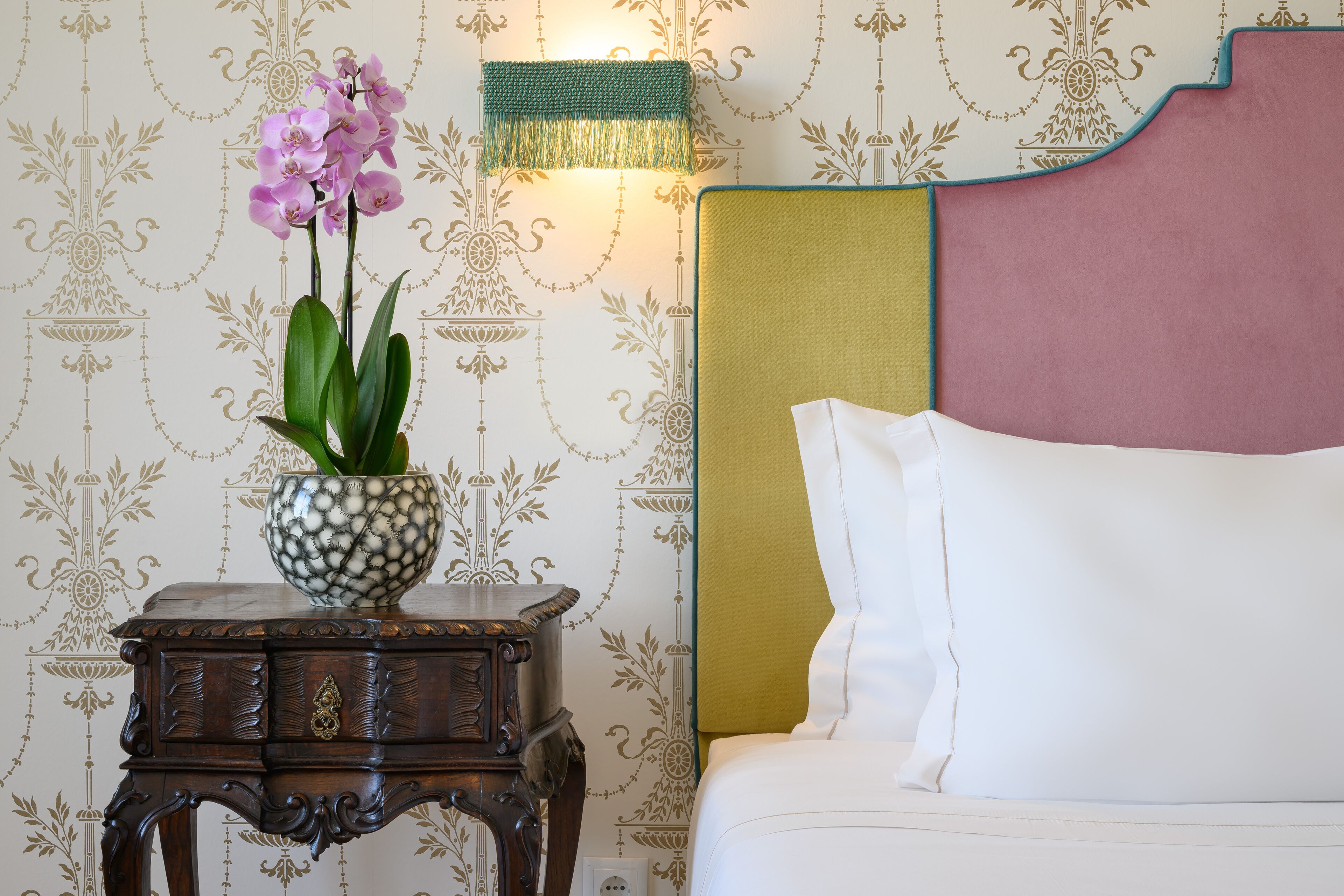 Un lit avec un oreiller blanc et une tête de lit aux couleurs rose et jaune. Une petite table de chevet en bois, sur laquelle se trouvent un vase à motifs et un abat-jour vert, est placée à côté du lit.