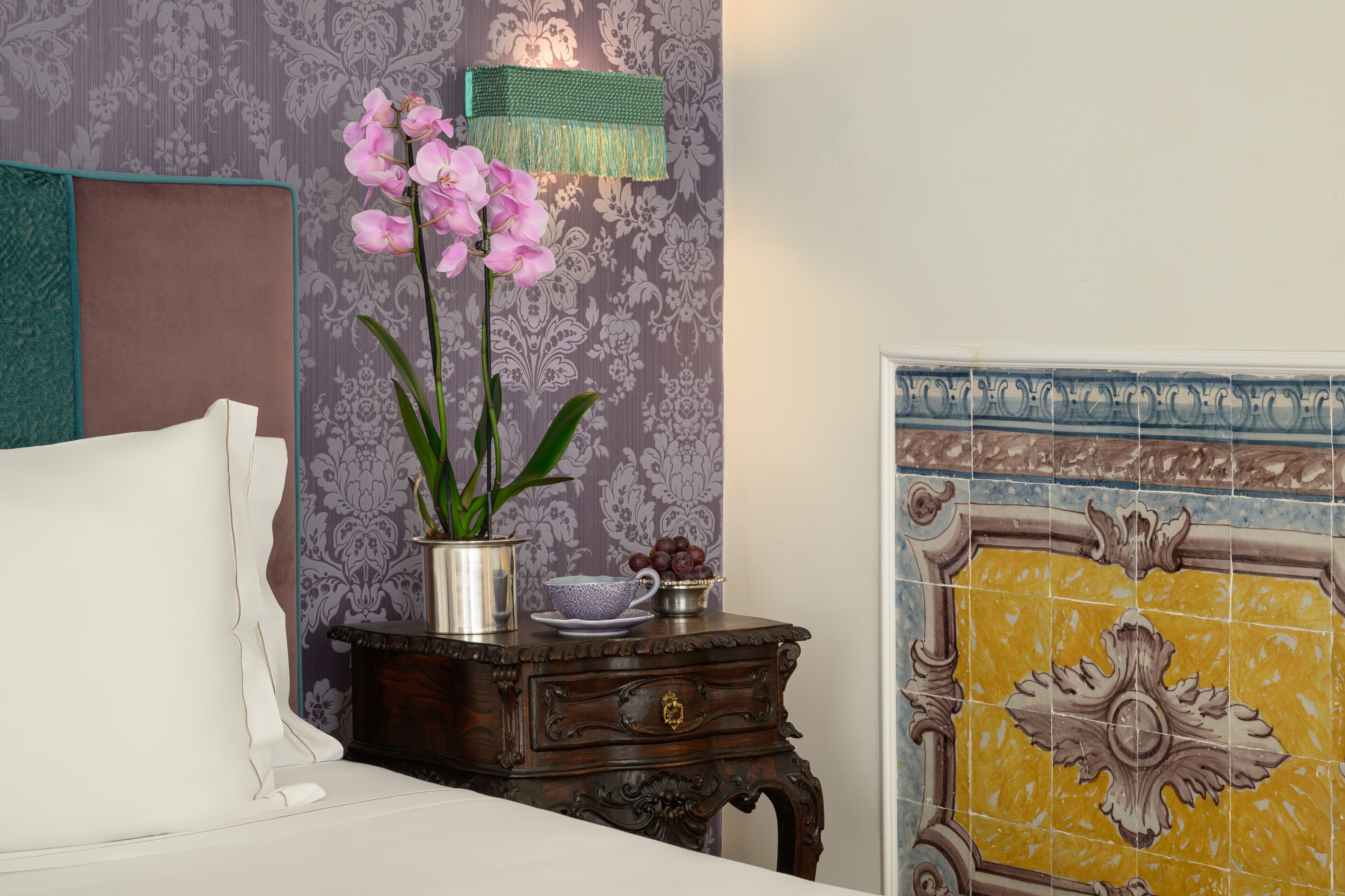 A neatly made bed with a white comforter, a bedside table with a vase of pink orchids, a metal pitcher, and a bowl of grapes.