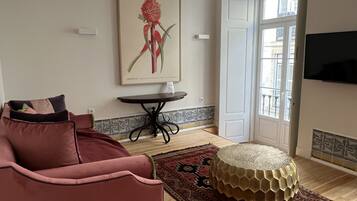 Ein Wohnzimmer mit einem roten Sofa, einem runden Hocker, einem kleinen Tisch und einem botanischen Druck im Bilderrahmen an der Wand.
