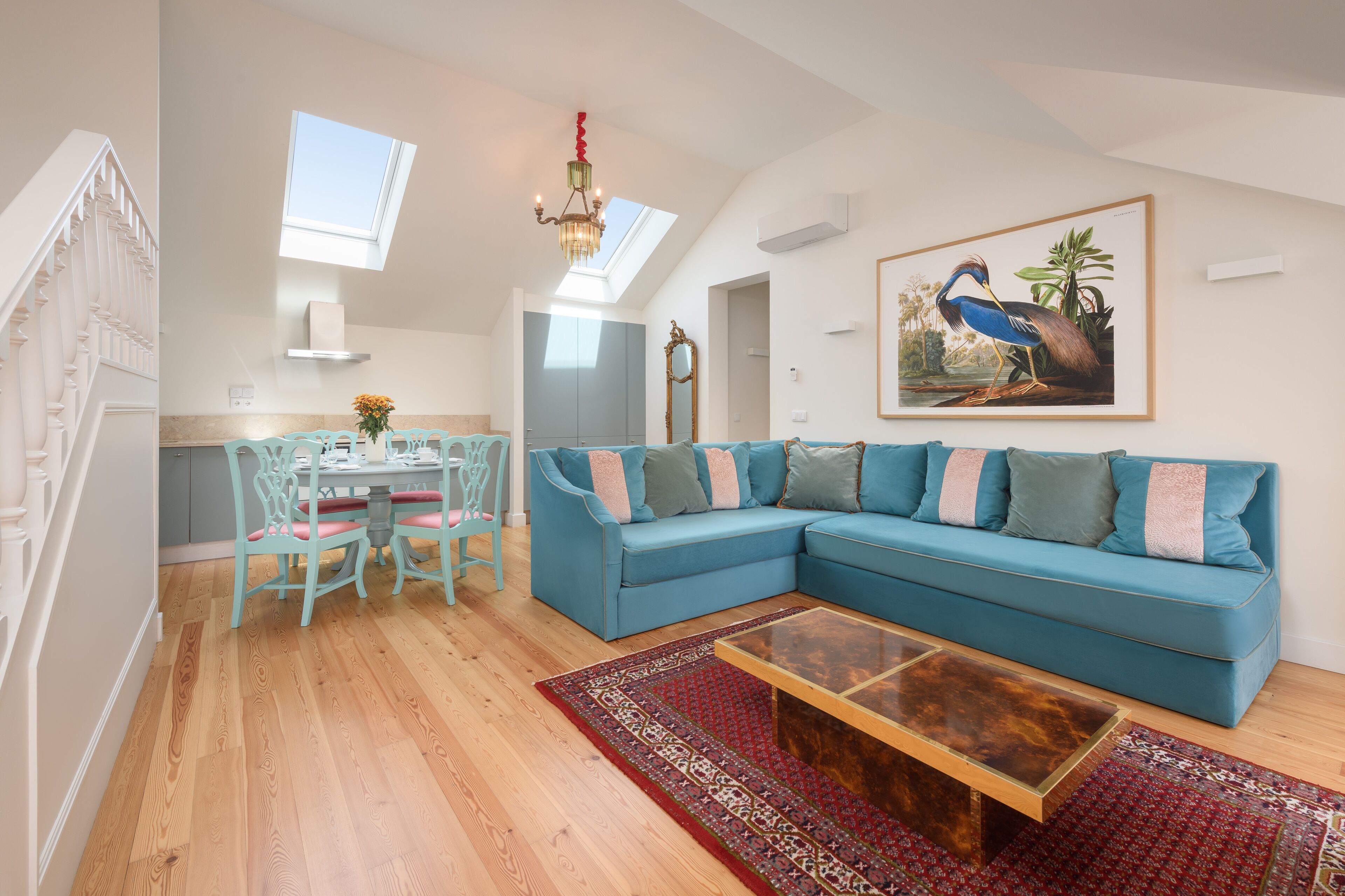 A spacious living area with a blue sofa, a dining table with chairs, a chandelier, and a large painting of a bird.