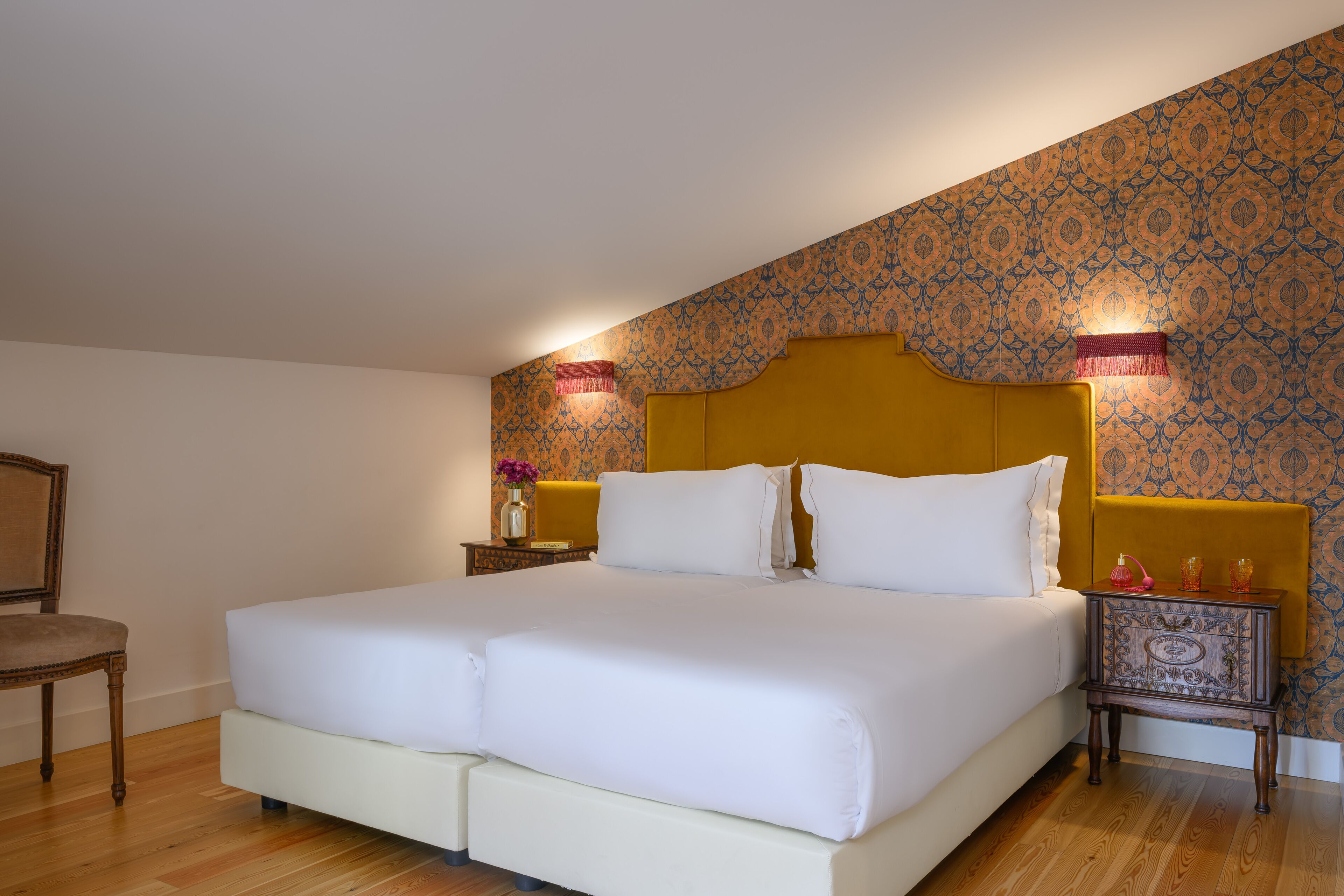 Una cama doble con ropa blanca, una mesita de noche de madera con un florero y dos velas, y un papel tapiz estampado.
