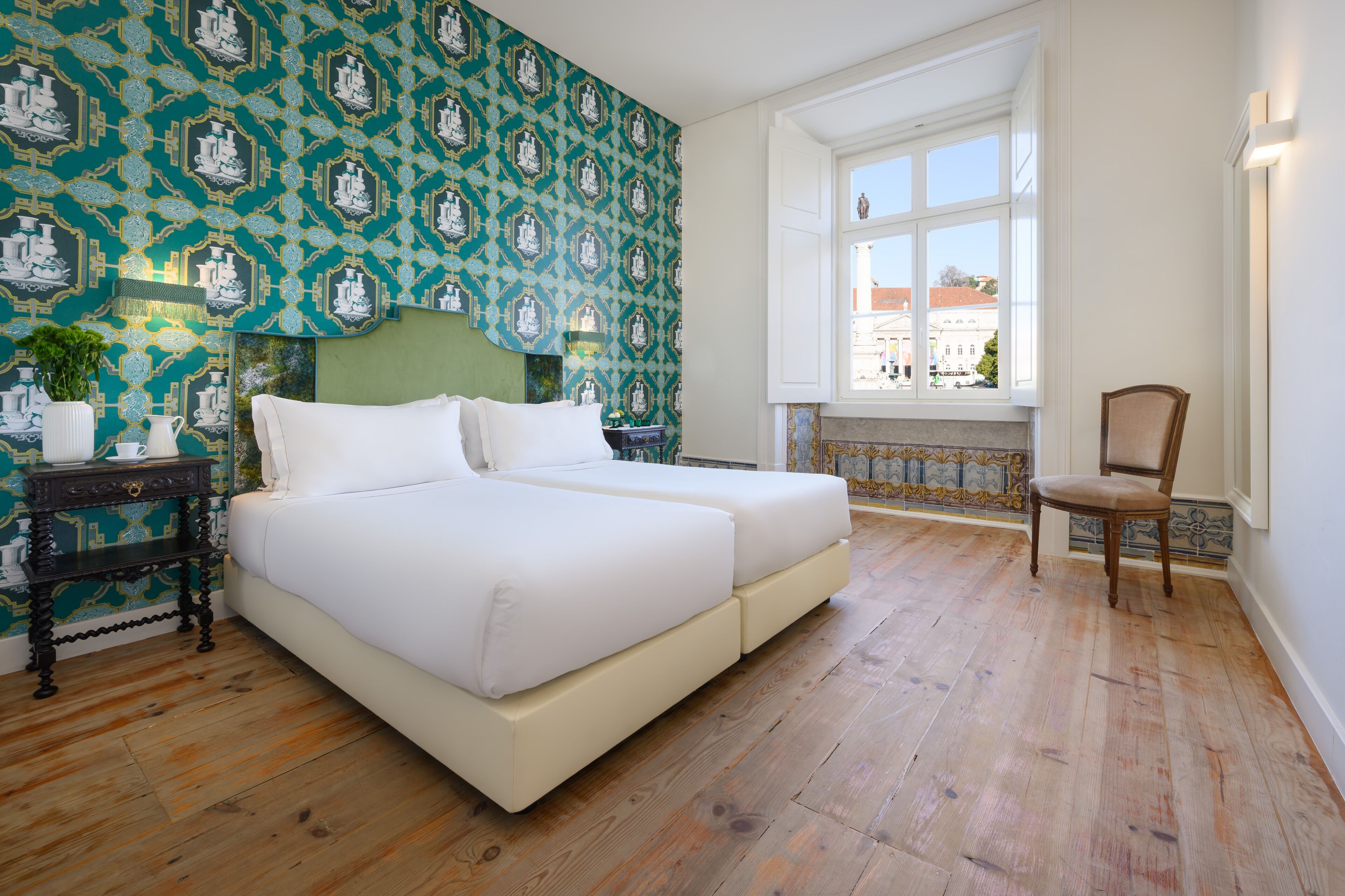 Ein Schlafzimmer mit einem großen Bett, einem Sessel, einem Nachttisch und einem Fenster mit Blick auf Gebäude.