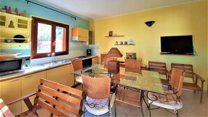 Eine kompakte Küche mit Mikrowelle, Herd und Spüle. Ein Essbereich mit Glastisch und Korbstühlen. Ein Fernseher ist an der Wand montiert.