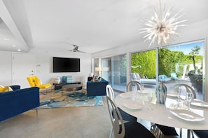 A modern living room with a large TV, blue sofas, a dining table set for four, and a balcony with greenery. - Joplin House - Poolside Paradise w/ Amazing Views! (Palm Springs)