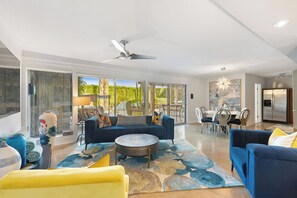 A spacious living room with a blue sofa, yellow armchair, and a round coffee table. There is a dining area with a table and chairs, and a kitchen with a refrigerator. - Joplin House - Poolside Paradise w/ Amazing Views! (Palm Springs)