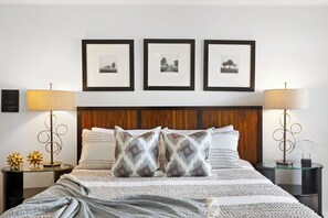 Een netjes ingerichte slaapkamer met een houten hoofdbord, een groot bed met gekleurde kussens, twee nachtkastjes met lampen en drie ingelijste foto’s aan de muur.