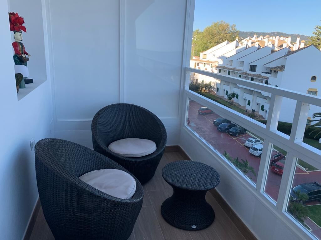 Un balcón con sillones de mimbre y una mesita redonda, con vista a una calle con coches aparcados y edificios blancos.