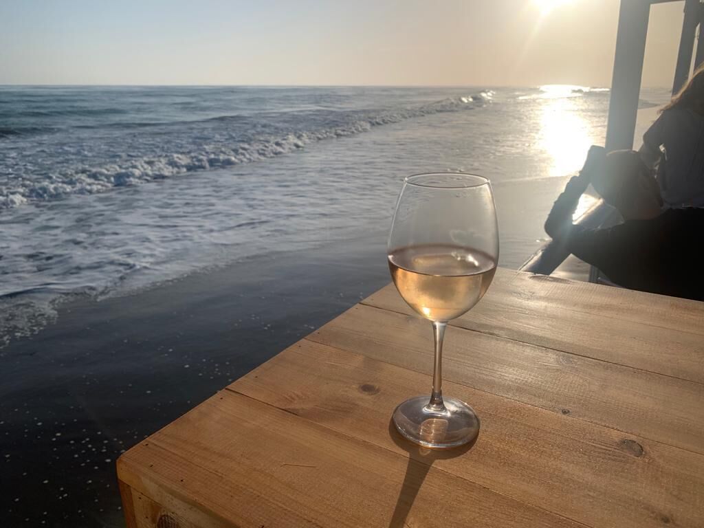 Un verre de vin sur une table en bois, près de la plage, avec l’océan en arrière-plan.