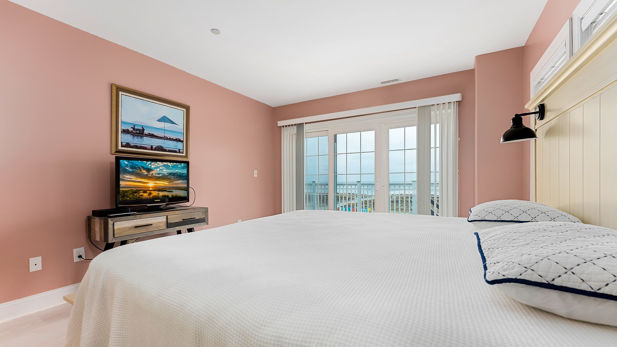 A bedroom with a large bed, a TV on a wooden stand, a painting, and a view of the sea through the window.