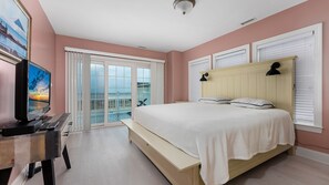 A bedroom with a large bed, a desk with a computer, a TV, and a balcony with a view. - 4br Beachfront Condo with Ocean Views and Pool (Wildwood Crest)