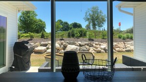 Vista a través de un ventanal a un patio con muro de piedra, una parrilla y mobiliario exterior.