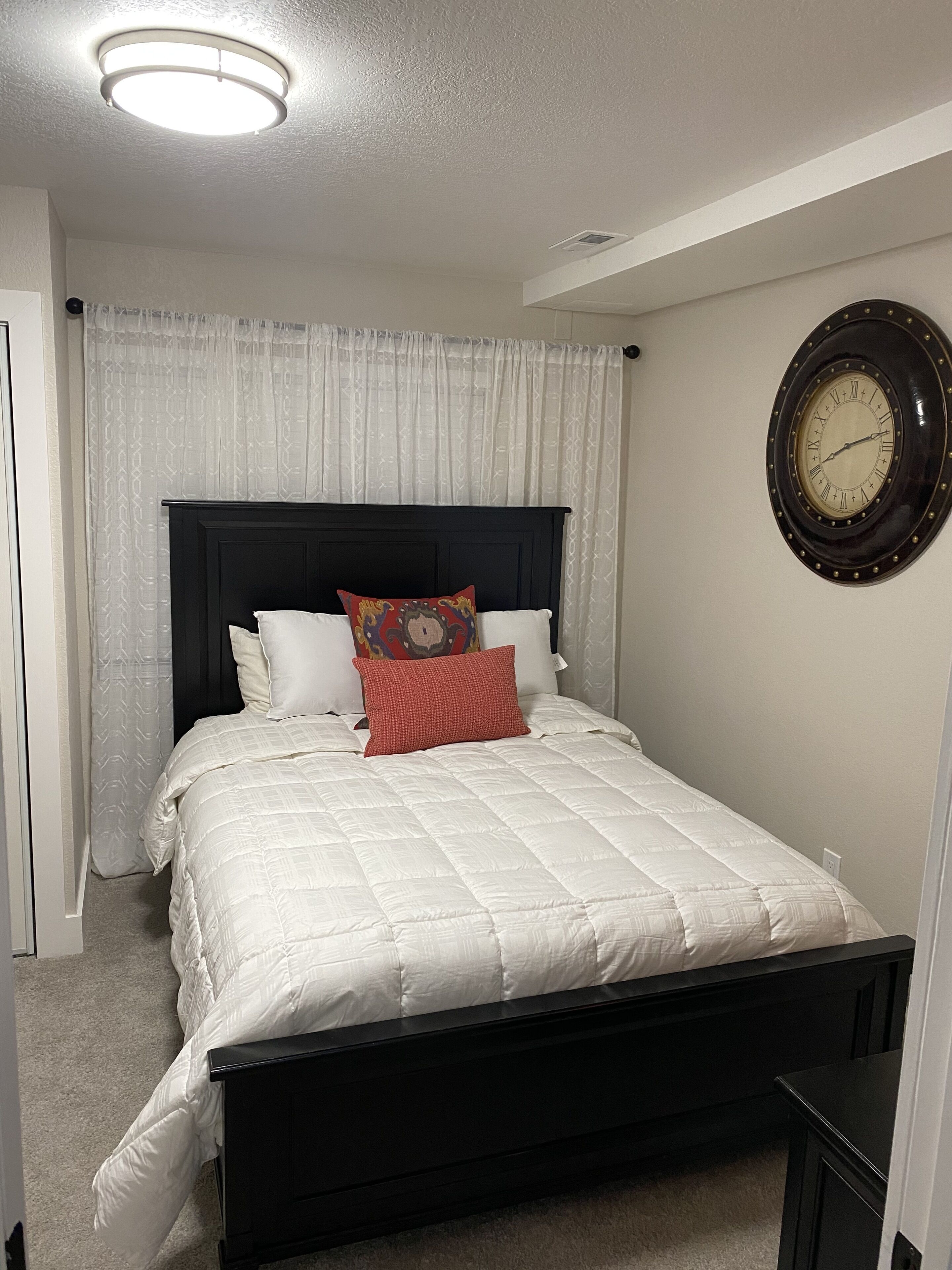 Una cama bien hecha con una colcha blanca y un cabecero negro, un reloj de pared redondo y una ventana con cortinas translúcidas.