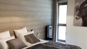 Quarto com teto de madeira, cama com roupa de cama branca e estampada, manta escura e televisão montada.