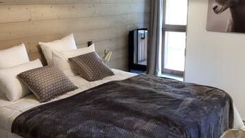 Une chambre avec un plafond en bois, un lit avec une literie blanche et à motifs, une couverture sombre et un téléviseur fixé au mur.