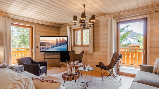 Interior acolhedor de uma cabana de madeira com televisão, assentos confortáveis e janelas amplas com vista panorâmica.