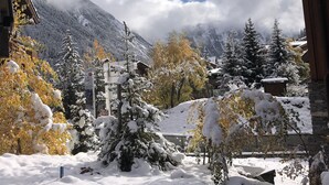Área externa nevada com um edifício de madeira, árvores cobertas de neve e uma montanha ao fundo.