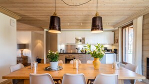 Cozinha moderna com armários de madeira, eletrodomésticos de aço inoxidável e uma área de jantar com mesa de madeira e cadeiras brancas.