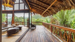 A wooden deck with a thatched roof, furnished with leather sofas and a wicker chair, overlooking a lush green forest. - Valley Stream Private Eden (St. Margaret's Village)