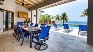 Een overdekt terras met eettafel en uitzicht op de oceaan, met een lange tafel met blauwe stoelen, een plafondventilator en een houten plafond.