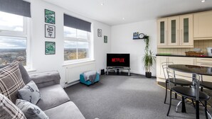A modern living room with a grey sofa, a small blue ottoman, a TV displaying 'NETFLIX', a dining area with a table and chairs, and a kitchen with cabinets. - Liquorice Suite (Lincoln)