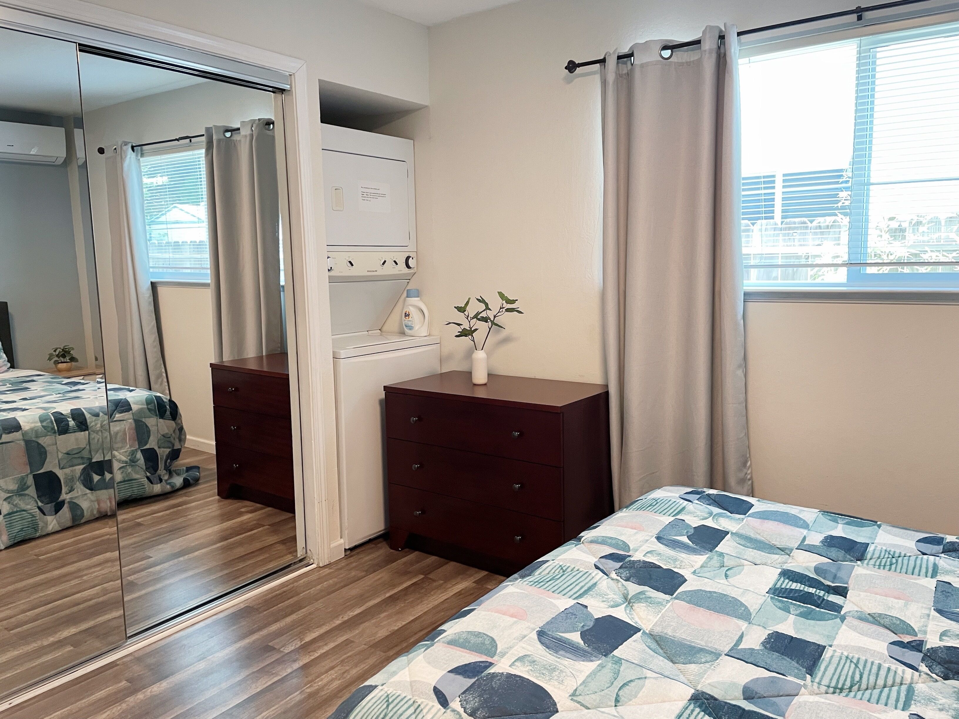 A bedroom with a wooden floor, a bed with a patterned bedspread, a dresser, a mirror, and a window with curtains.