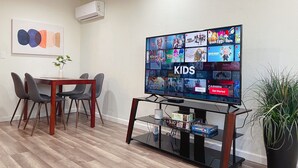 A modern living room with a flat-screen TV displaying a children's channel, a dining area with a table and chairs, and a potted plant. - 💎Cozy 1B/1B in a prime location • 🌲AC/ 1000mb wifi/ Washer, dryer/ more… (Sunnyvale)