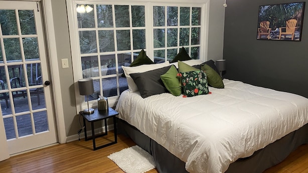 A bedroom with a large bed, a nightstand, a lamp, and a window with a view of the outdoors. - Suburb in the City (Atlanta)