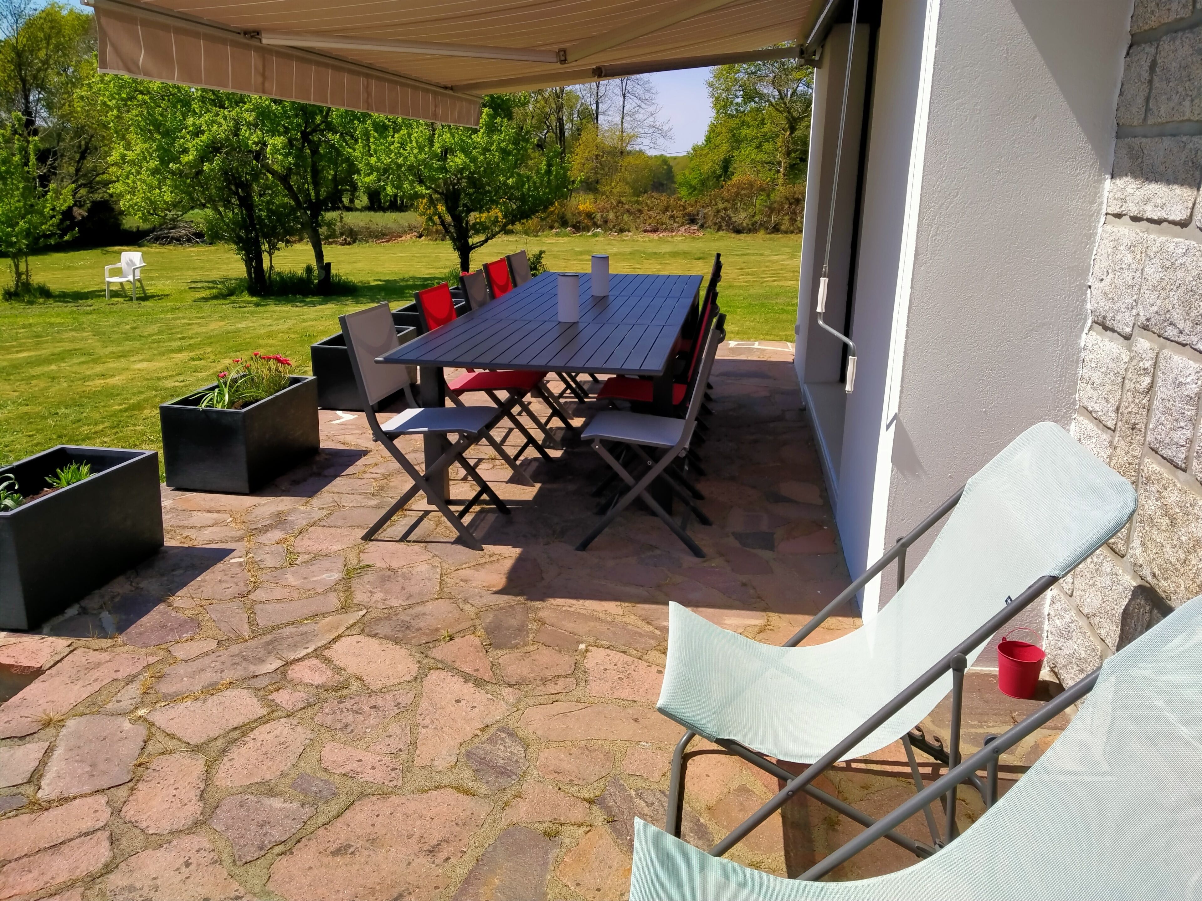 En udendørs terrasse med et bord og stole, omgivet af stenbelægning og krukker med planter.