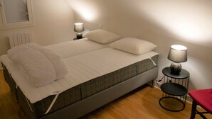 Una cama bien tendida con ropa de cama blanca, una mesita de noche pequeña con una lámpara y una silla roja en una habitación sencilla con suelo de madera.