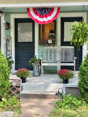 A front porch with a black door, a welcome sign, a bench, and potted plants.
