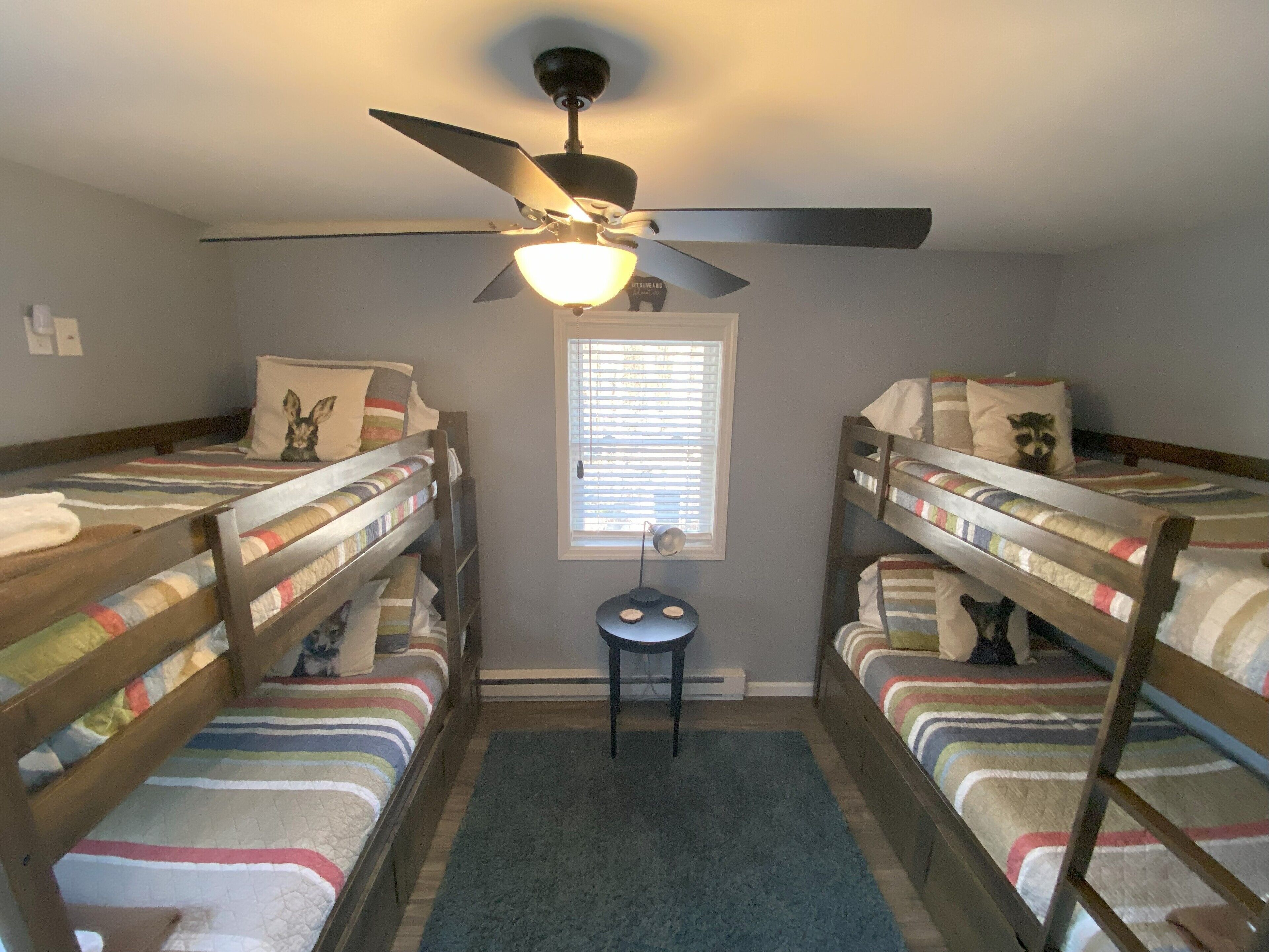 A room with two bunk beds, a ceiling fan, a small table, and a window with blinds.