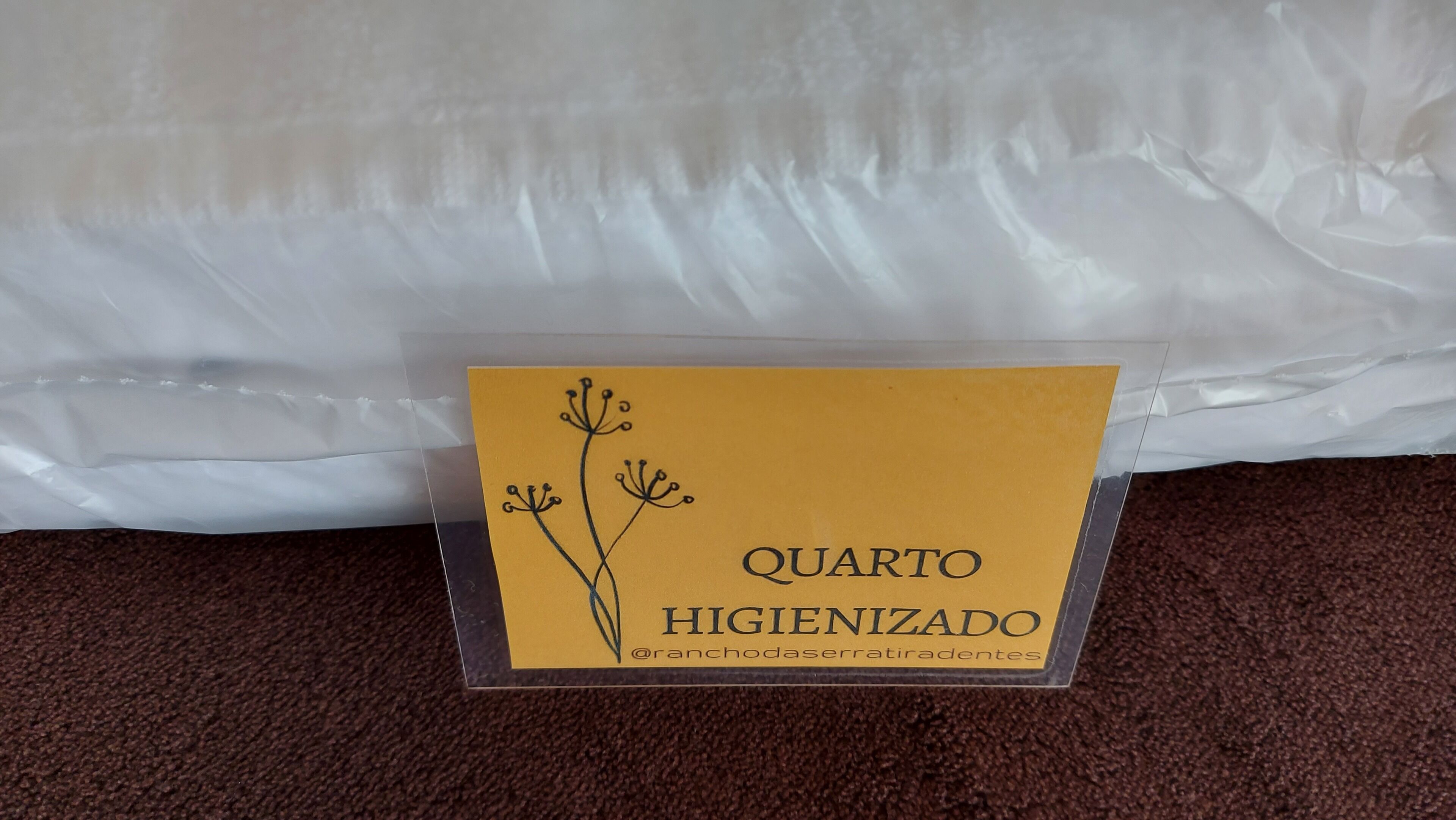 Tarjeta de plástico transparente con el texto "QUARTO HIGIENIZADO" y un diseño floral, sobre una alfombra marrón sobre fondo blanco.