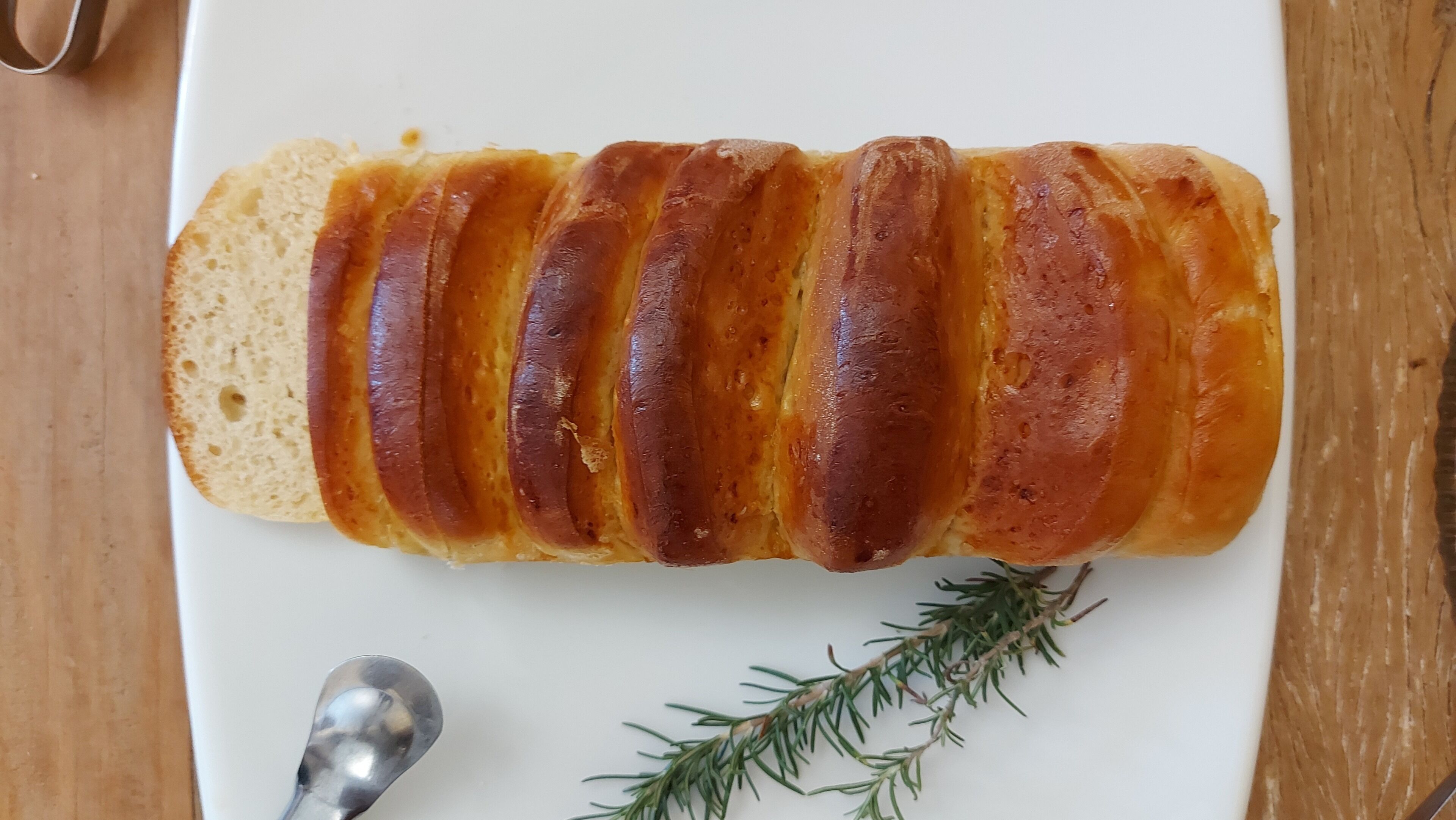 A loaf of bread with a golden-brown crust, placed on a white plate with a sprig of rosemary and a spoon beside it.