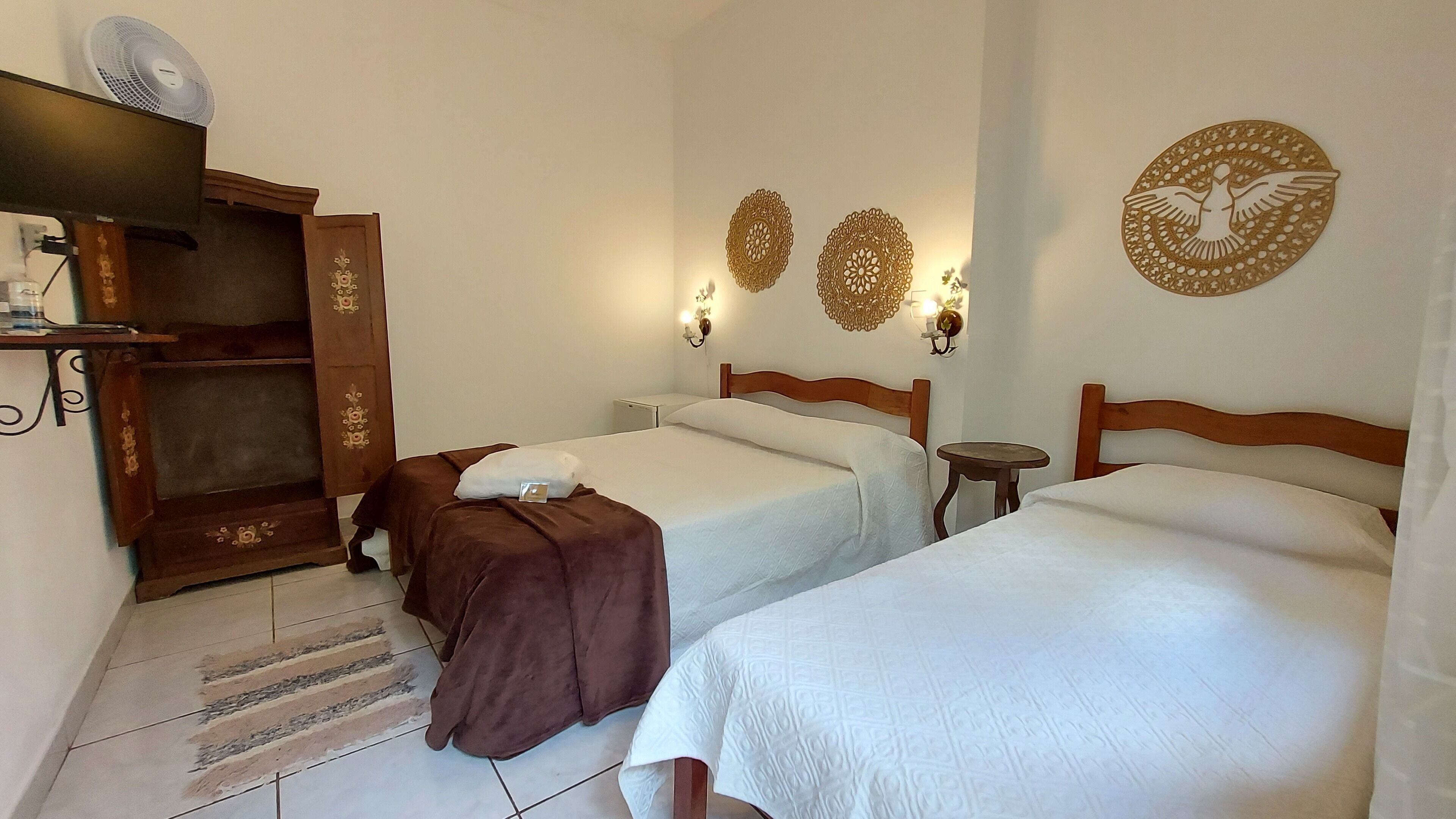 A hotel room with two beds, a wooden wardrobe, a small table, and decorative wall sconces.