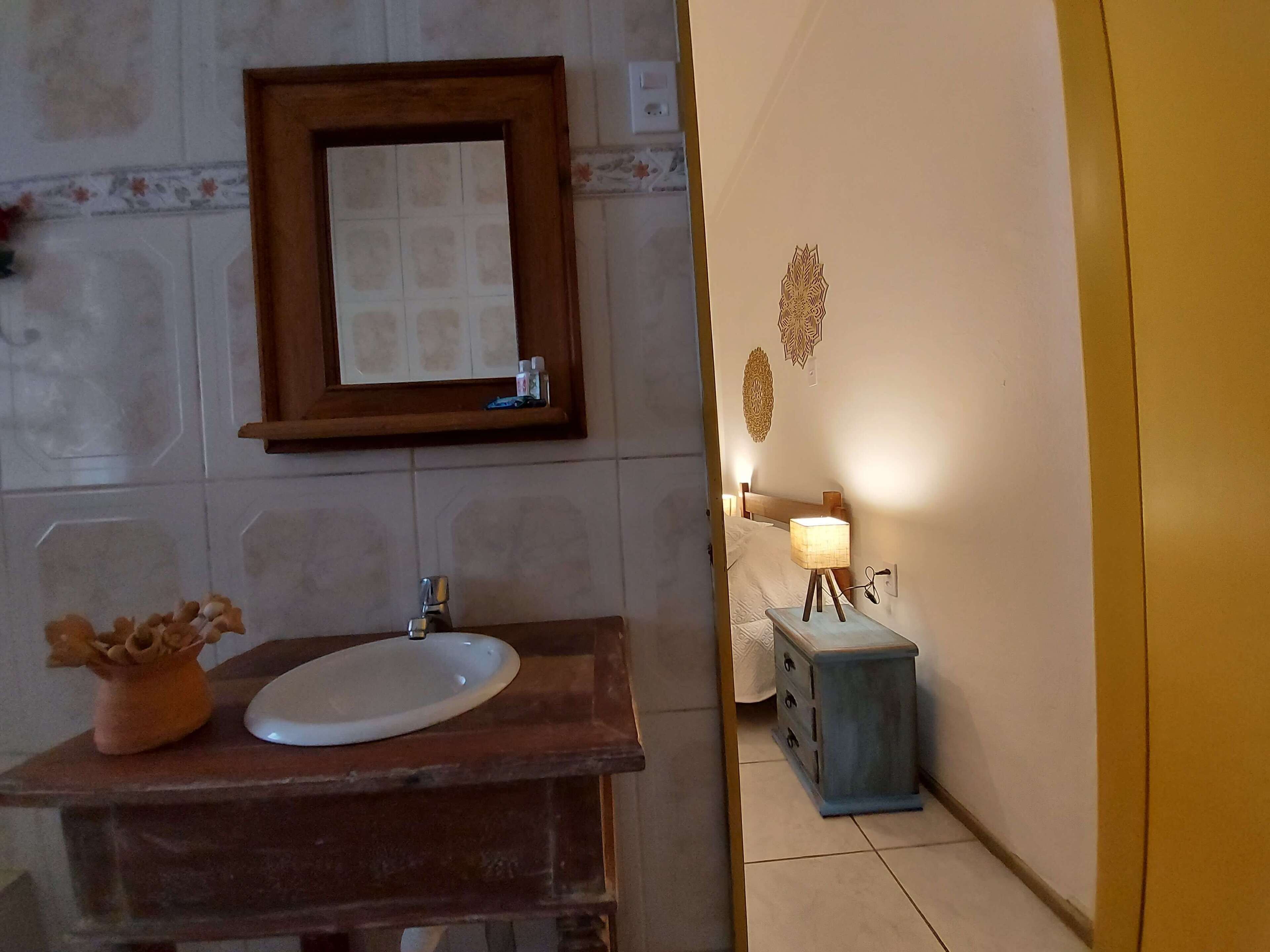 A bathroom with a wooden vanity, a white sink, a mirror, and a bedside table with a lamp.