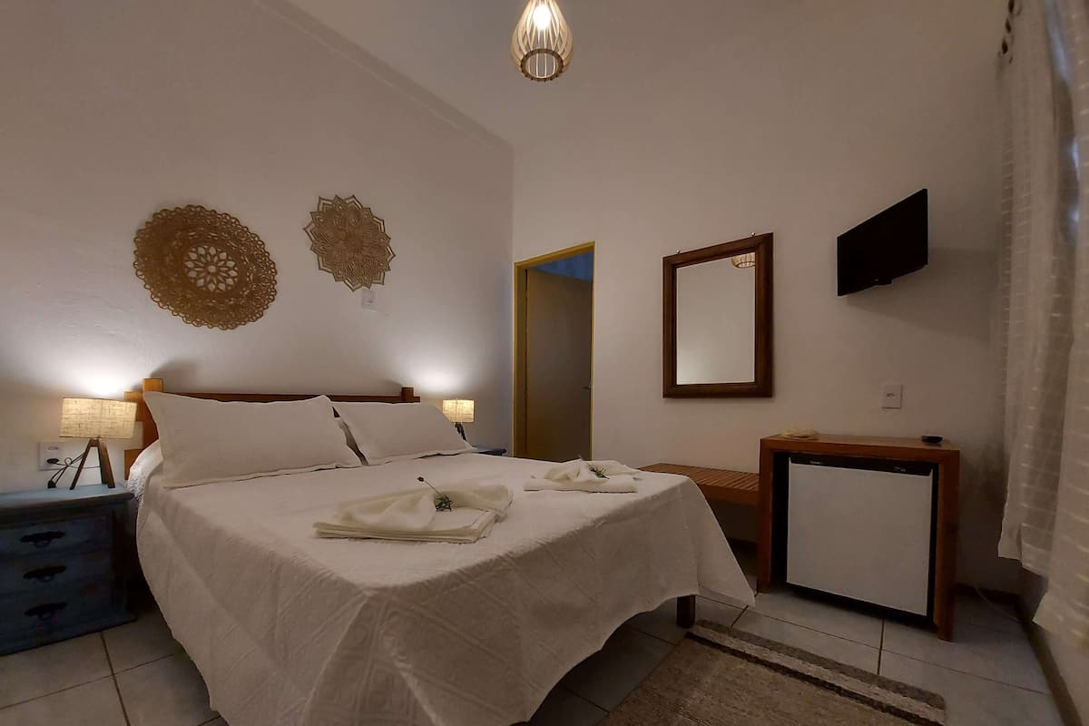 Una cama bien hecha con una colcha blanca y almohadas, una mesita de noche con una lámpara, un televisor montado en la pared y un espejo.