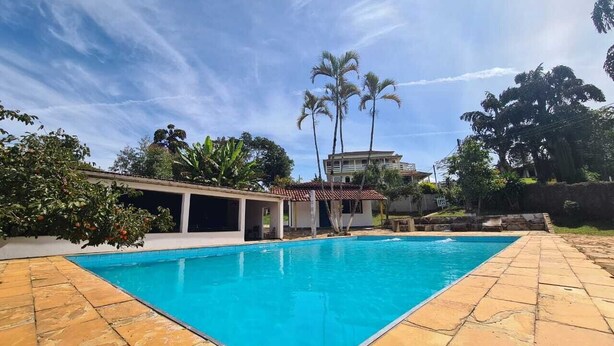 A rectangular swimming pool with a tiled deck, surrounded by palm trees and a building with a tiled roof in the background. - Rancho da Serra Pousada (Tiradentes)