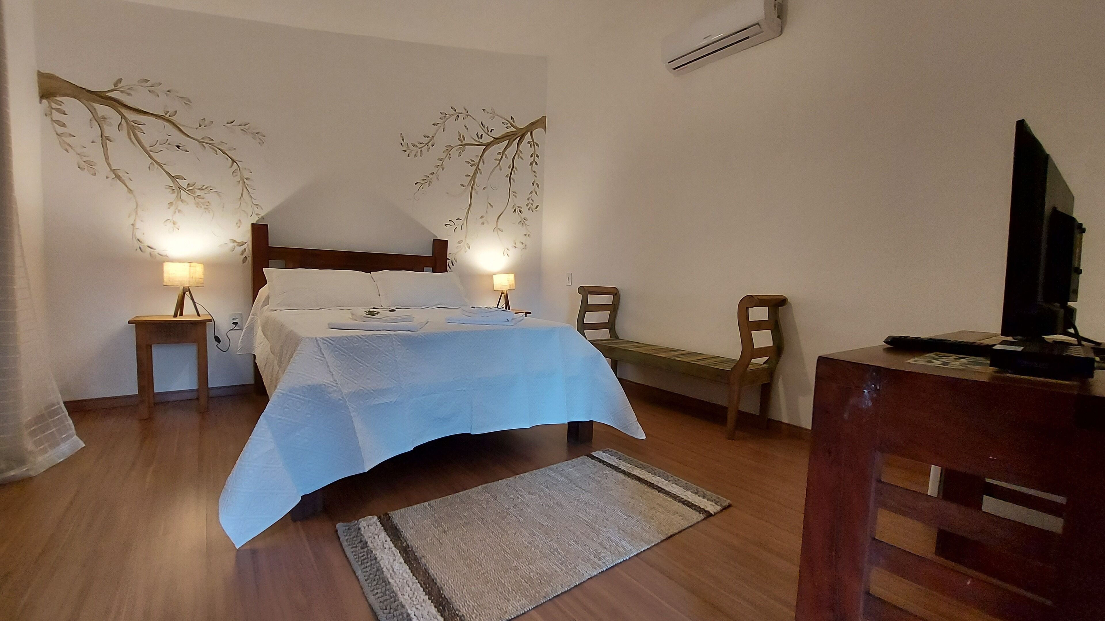 A bedroom with a bed, bedside lamps, a wooden chair, a desk with a computer, and a wall-mounted air conditioner.