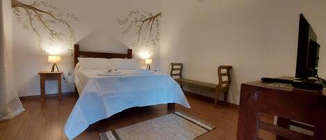 A bedroom with a bed, bedside lamps, a wooden chair, a desk with a computer, and a wall-mounted air conditioner.