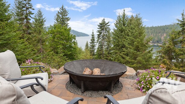 A patio with a stone fire pit surrounded by chairs, overlooking a lake and forested hills. - Luxurious Echo Bay Retreat—Hot Tub, Fire Pit, Ping Pong, Chess, Bikes & Stunning Views Await! (Harrison)