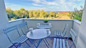 Un balcon avec une table et des chaises, un pot de fleurs et une vue sur des arbres et une côte lointaine.