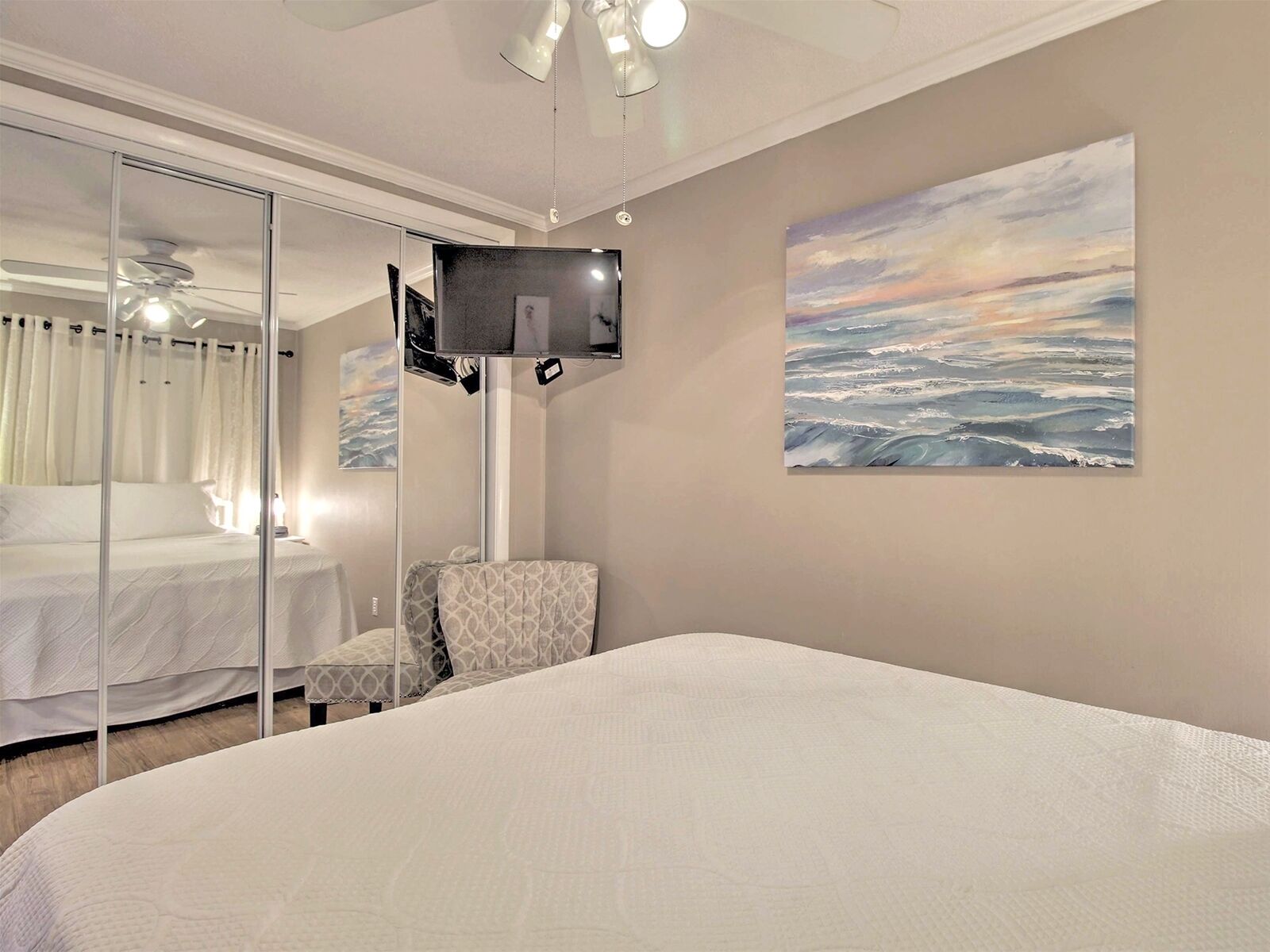A hotel room with a bed, a chair, a TV mounted on the wall, and a painting of a seascape.