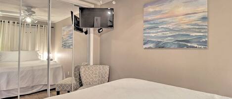 A hotel room with a bed, a chair, a TV mounted on the wall, and a painting of a seascape.