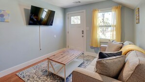 Una sala de estar con un televisor de pantalla plana montado en la pared, una mesa de centro de madera, un sofá con cojines y una ventana con cortinas amarillas.