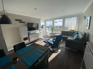 A modern living room with a flat-screen TV, a sofa, a dining table with chairs, and large windows offering a view of the city. - Skriien Mö (Sylt)
